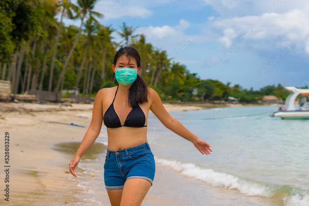 Fototapeta premium use of medical face mask in public places- young attractive Asian Korean woman enjoying beach holidays in bikini and protective facial mask in prevention vs virus infection