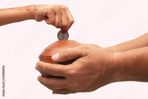 Hands Holding Indian penny bank made with mud clay and Child hand adding coin into the earthen pot or terracotta piggy bank on Isolated background - Concept showing of saving, investement or donating. photo