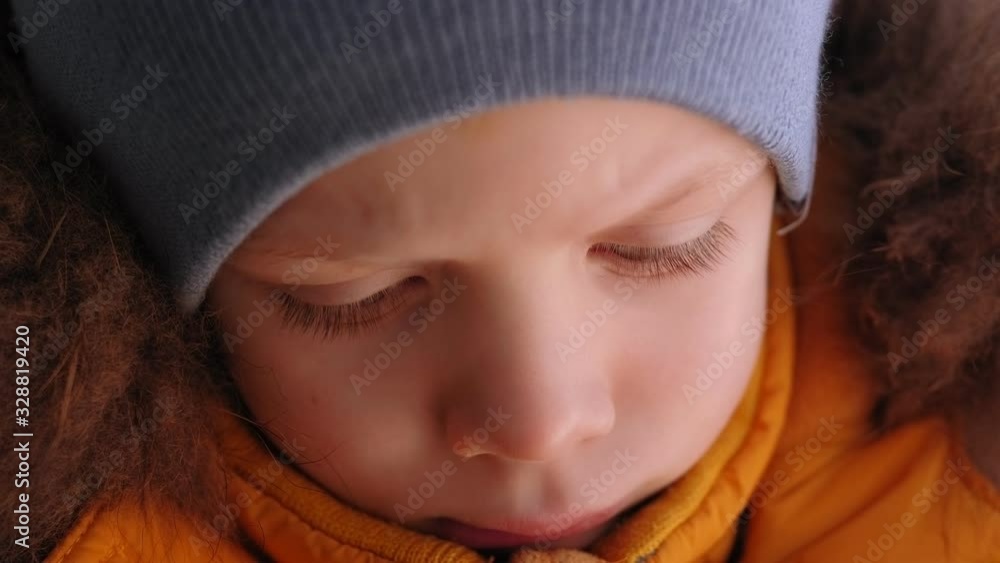 Close-up of the face of a small sad boy in a warm winter jacket with a fur hood and a hat, he says something upset. Close-up of the face of a small sad boy in a warm winter jacket with a fur hood and a hat, he says something upset.