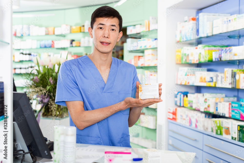 Portrait of chinese man pharmacist who is standing on his work place near cashbox in apothecary.