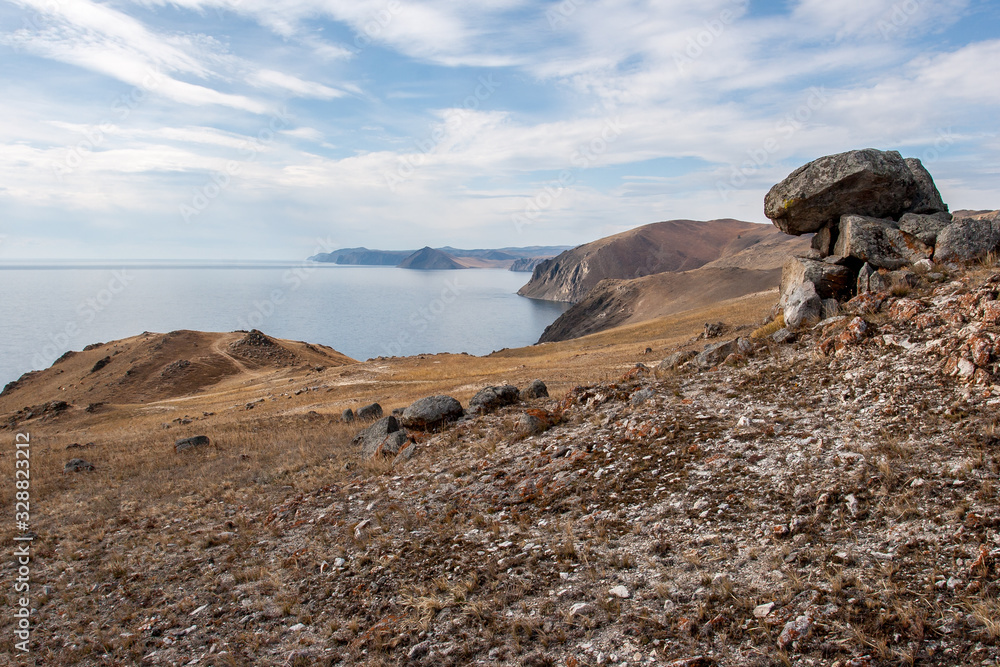 The shore of Lake Baikal with stones in the foreground. High rocky shore. There are clouds on the blue sky. The lake is calm.