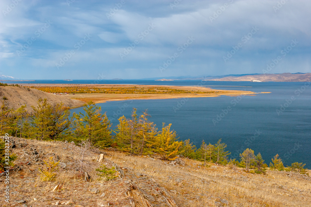 Autumn landscape overlooking Lake Baikal. Yellow trees and blue water. Stones on the ground. Clouds in the sky and rain in the distance. Horizontal. Autumn landscape overlooking Lake Baikal. Yellow trees and blue water. Stones on the ground. Clouds in the sky and rain in the distance. Horizontal.