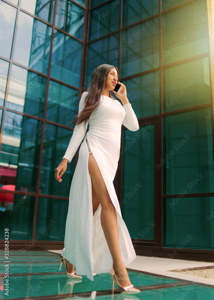 Fashion afro woman in a luxurious white dress talking on phone while walking against building with blue windows outdoor. Fashion afro woman in a luxurious white dress talking on phone while walking against building with blue windows outdoor.