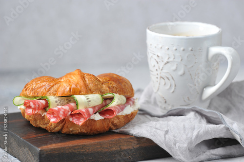 Croissant stuffed with salami, fresh cucumber and cottage cheese. Next to the croissant is a cup of coffee. Close-up. Light background. 