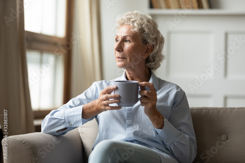 Принти на полотні Pensive senior 60s woman sit rest on couch in living room drink tea or coffee lo