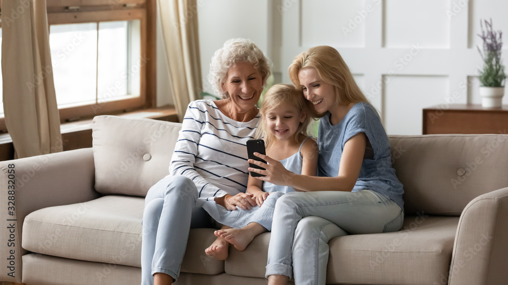 Smiling three generations of women sit on couch in living room have fun use smartphone together, happy cute little girl with young mother and grandmother watch funny video on cellphone relax at home Smiling three generations of women sit on couch in living room have fun use smartphone together, happy cute little girl with young mother and grandmother watch funny video on cellphone relax at home