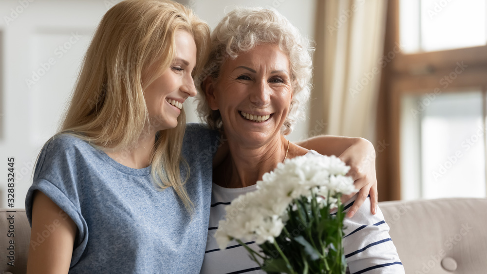 Portrait of overjoyed mature mother and grownup daughter sit on couch at home celebrate birthday together, loving caring adult girl child congratulating senior mom presenting flowers bouquet Portrait of overjoyed mature mother and grownup daughter sit on couch at home celebrate birthday together, loving caring adult girl child congratulating senior mom presenting flowers bouquet