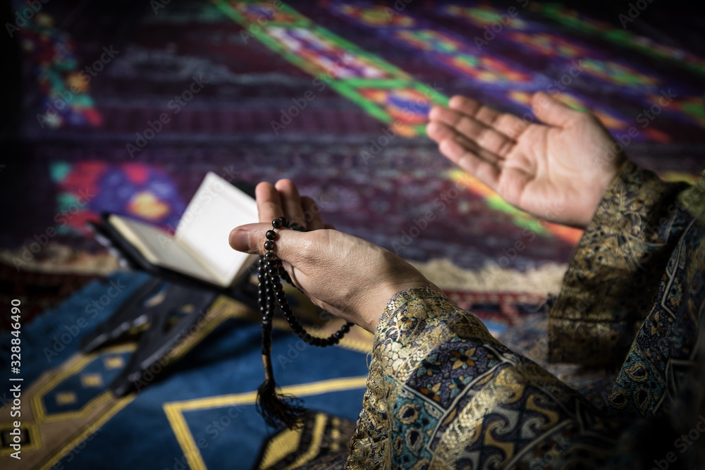 Obraz premium Muslim woman praying for Allah muslim god at room near window. Hands of muslim woman on the carpet praying in traditional wearing clothes, Woman in Hijab, Carpet of Kaaba, Selective focus, toned
