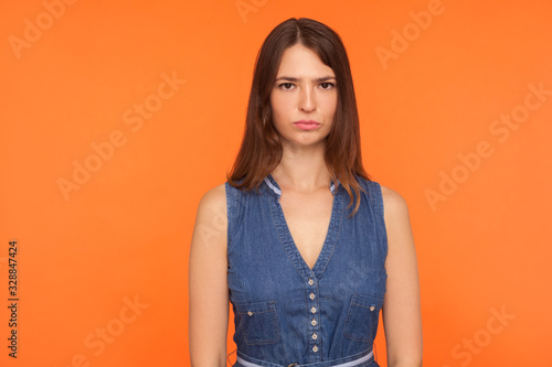 Unhappy brunette woman in denim dress looking at camera with depressed gloomy face expression, feeling melancholy dismal, worried about problems. indoor studio shot isolated on orange background