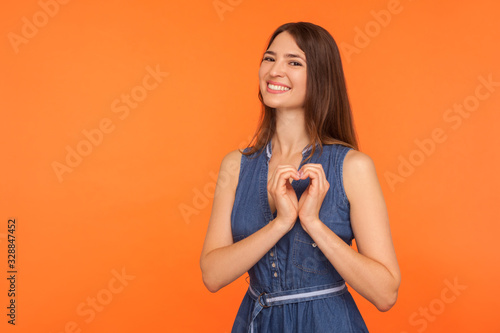 Cheerful pretty woman in dress making heart shape with hands and smiling kindly, showing charity symbol, declaration of love, expressing emotions. indoor studio shot isolated on orange background