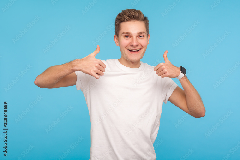 Well done! Portrait of happy enthusiastic man in casual white t-shirt showing thumbs up and smiling friendly, being delighted satisfied with result. indoor studio shot isolated on blue background - obrazy, fototapety, plakaty 