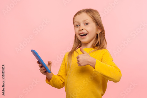 Best app for kids. Happy excited little girl holding smartphone and showing thumb up, like gesture to appreciate video game, satisfied with easy mobile app. studio shot isolated on pink background photo