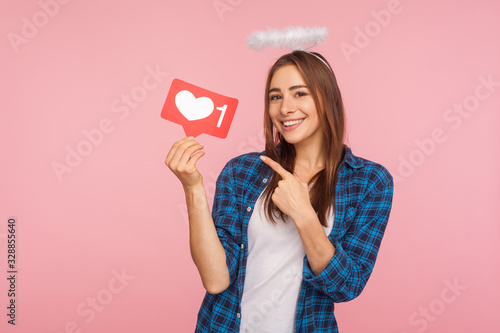 Portrait of happy charming girl with angelic halo smiling and pointing at social media like icon, heart button to enjoy trendy interesting content. indoor studio shot isolated on pink background