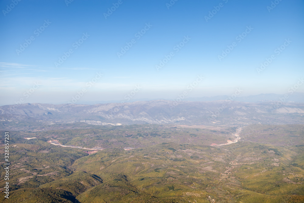 Areal view of a curvy highway road going through the valleys and forest with background mountains and blue sky