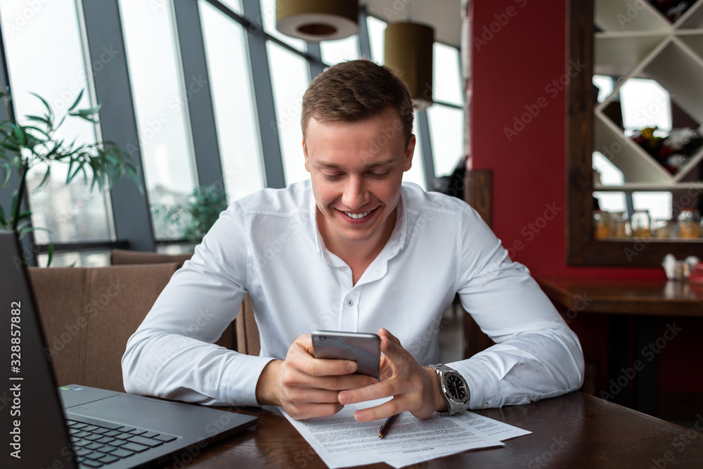 Young businessman wearing a white fashion shirt chatting and talking business on a mobile phone in a cafe with a laptop and documents. Freelance and selfemployment concept. Distance job. Smiling Young businessman wearing a white fashion shirt chatting and talking business on a mobile phone in a cafe with a laptop and documents. Freelance and selfemployment concept. Distance job. Smiling