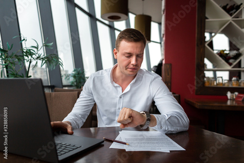 Young businessman wearing a white fashion shirt sitting in a cafe checking time on watches waiting for business partner with documents and laptop. Freelance and selfemployment concept. Distance job.