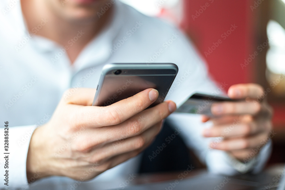 Closeup of hands of young businessman wearing a white fashion shirt with a mobile phone and a credit card making payment online in a cafe. Freelance and selfemployment concept. Distance job.