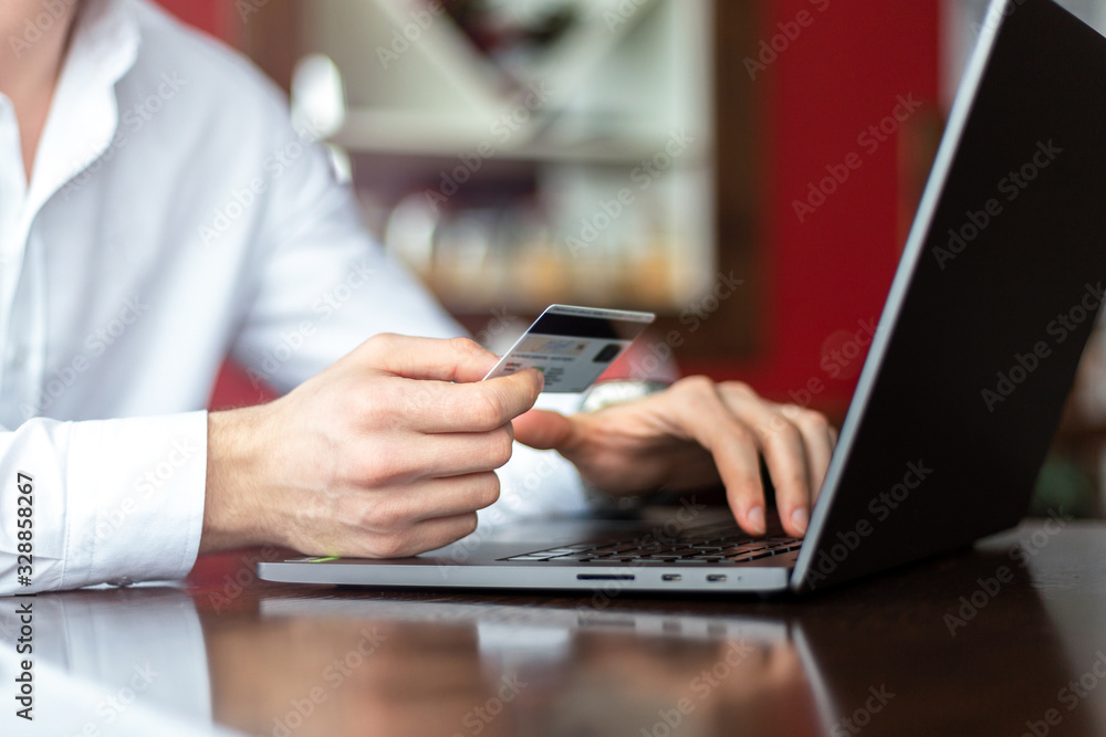 Closeup of hands of young businessman wearing a white fashion shirt with a credit card and laptop making payment online in a cafe. Freelance and selfemployment concept. Distance job. Closeup of hands of young businessman wearing a white fashion shirt with a credit card and laptop making payment online in a cafe. Freelance and selfemployment concept. Distance job.