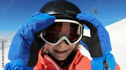 Smiling child skiing and waving hand on snow mountain and blue sky background. Happy boy enjoying winter vacation on ski slopes in sunny day. Having fun with outdoors activity, sport