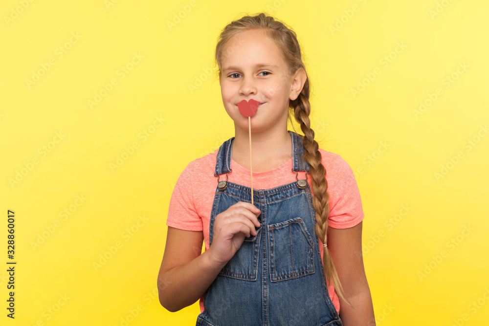 Portrait of charming happy little girl with braid in denim overalls covering mouth with red paper lips on stick and smiling at camera, symbol of kiss. indoor studio shot isolated on yellow background Portrait of charming happy little girl with braid in denim overalls covering mouth with red paper lips on stick and smiling at camera, symbol of kiss. indoor studio shot isolated on yellow background