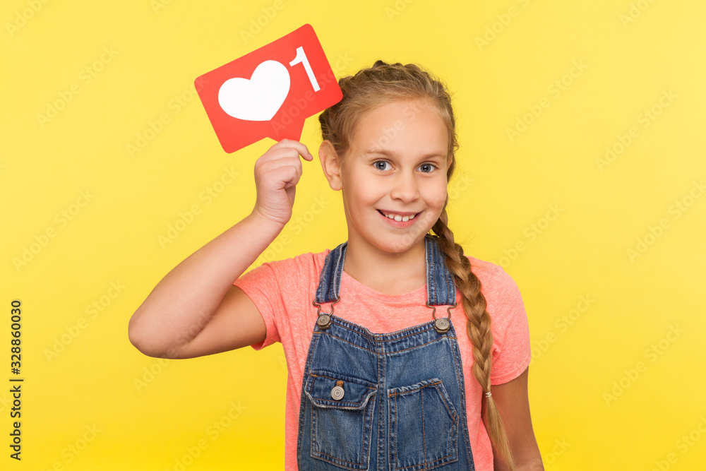 Like and comment for child blogging. Portrait of happy little girl in denim overalls holding social network heart button, follower notification symbol, popular internet content. studio shot isolated Like and comment for child blogging. Portrait of happy little girl in denim overalls holding social network heart button, follower notification symbol, popular internet content. studio shot isolated