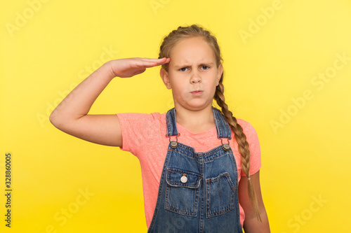 Yes sir! Portrait of responsible obedient little girl with braid in denim overalls saluting and looking at camera with respect, patriotic child. indoor studio shot isolated on yellow background