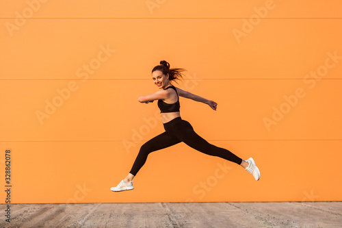 Positive lively vivid girl with fit body in tight sportswear jumping, running and smiling full of energy, healthy sport lifestyle. outdoor studio shot isolated on orange background, advertising area
