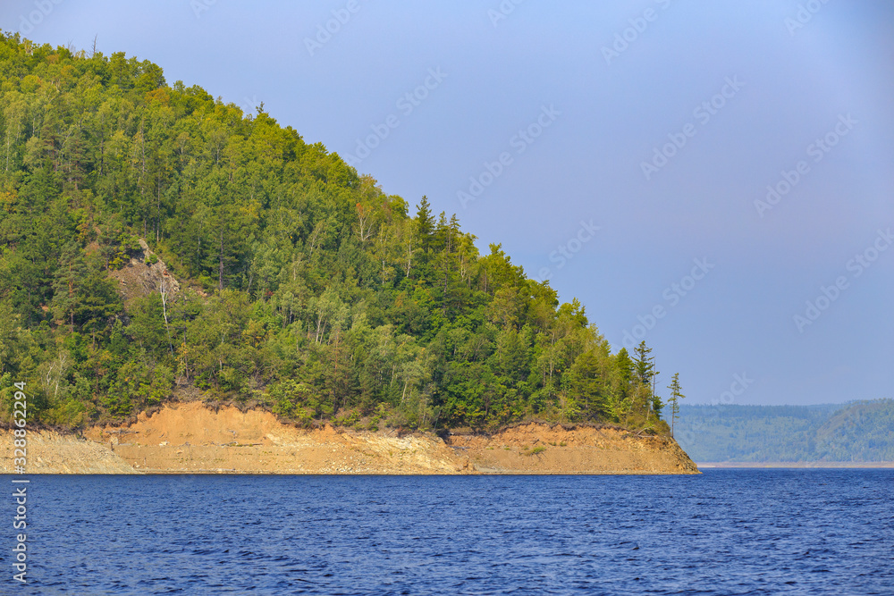 Beautiful landscape. Zeya reservoir, Amur region. A beautiful rocky shore with tall green trees goes into the calm blue waters of the reservoir.
