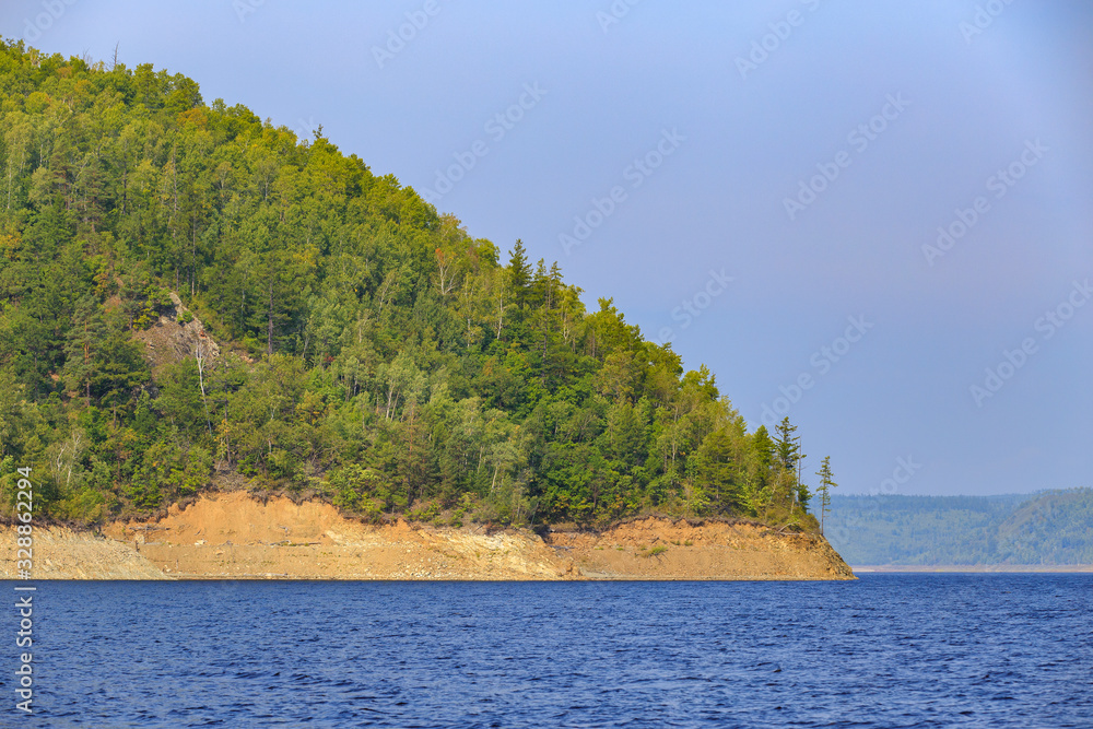 Obraz premium Beautiful landscape. Zeya reservoir, Amur region. A beautiful rocky shore with tall green trees goes into the calm blue waters of the reservoir.