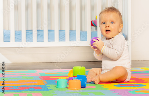 Little toddler baby boy sitting on children carpet near crib building pyramid of blocks looking at camera