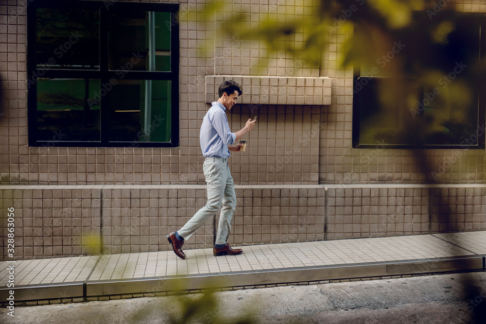 Happy Young Businessman in Casual wear Using or reading Mobile Phone while Walking by the Urban Building. Lifestyle of Modern People. Side View. Full Length Happy Young Businessman in Casual wear Using or reading Mobile Phone while Walking by the Urban Building. Lifestyle of Modern People. Side View. Full Length
