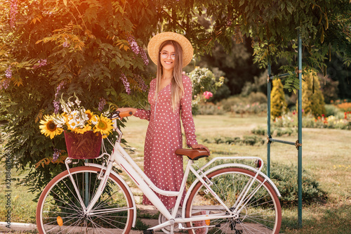 Young pleased woman in straw hat and glasses with bike in summer garden