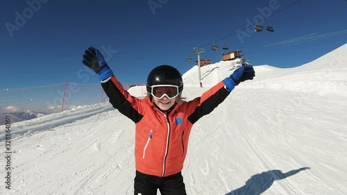 Smiling child skiing and waving hand on snow mountain and blue sky background. Happy boy enjoying winter vacation on ski slopes in sunny day. Having fun with outdoors activity, sport