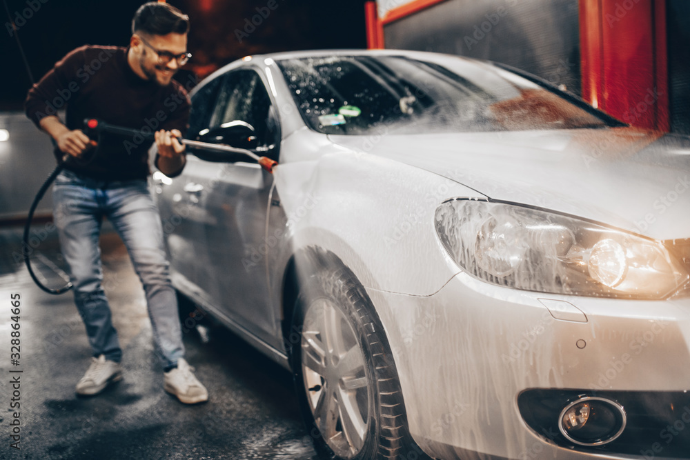 Young man washing his car in the evening at car wash station using high pressure water.