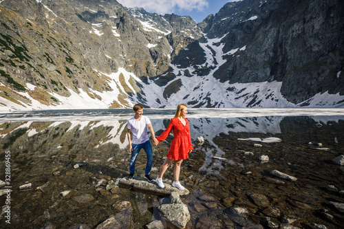 a young couple, a girl in a red dress, a man in a white T-shirt and blue pants stand on a background of mountains near the lake in the park