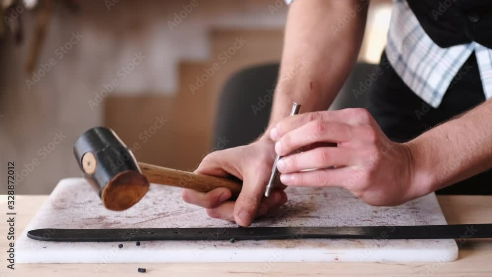 A leather craftsman is making holes in a hand-made belt. The tanner is using a mallet and a hole punch. A leather craftsman is making holes in a hand-made belt. The tanner is using a mallet and a hole punch.