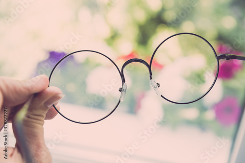 Female hand holds round glasses against the background of flowers and a window, the direction of view through the glasses