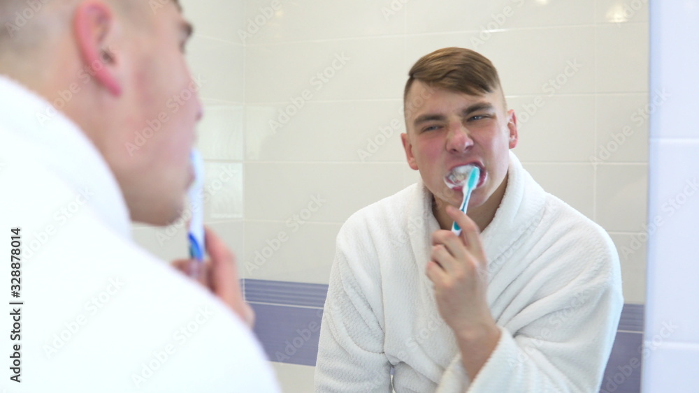 A young man is actively brushing his teeth. A man in a white bathrobe in front of a bathroom mirror. View through the mirror