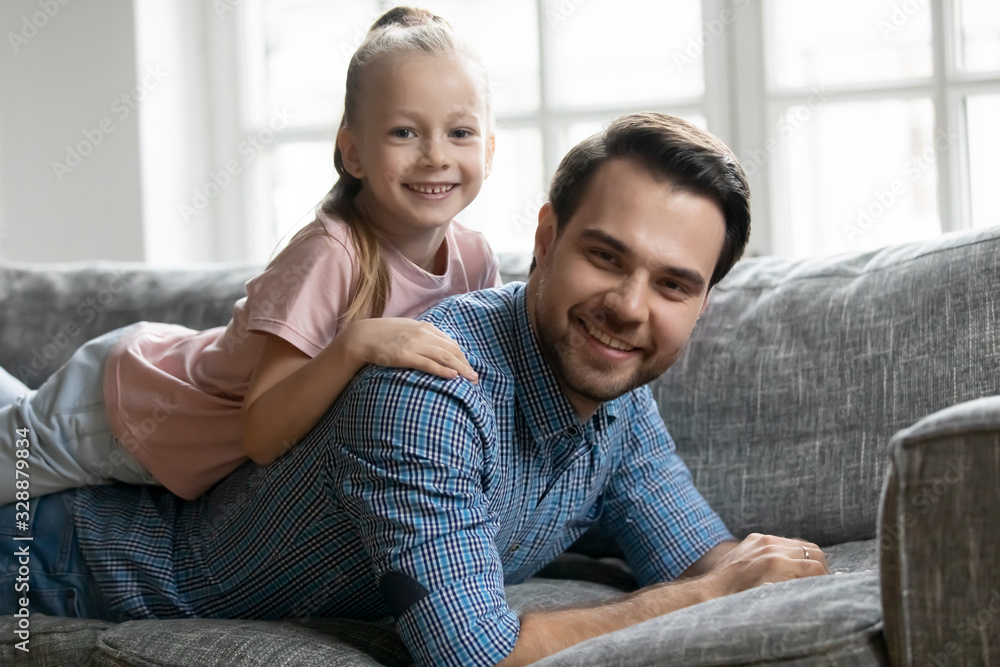 Portrait of smiling pleasant young father lying on comfy sofa, holding cute little blonde kid daughter on back. Affectionate two generations family enjoying weekend time, resting on couch at home. Portrait of smiling pleasant young father lying on comfy sofa, holding cute little blonde kid daughter on back. Affectionate two generations family enjoying weekend time, resting on couch at home.