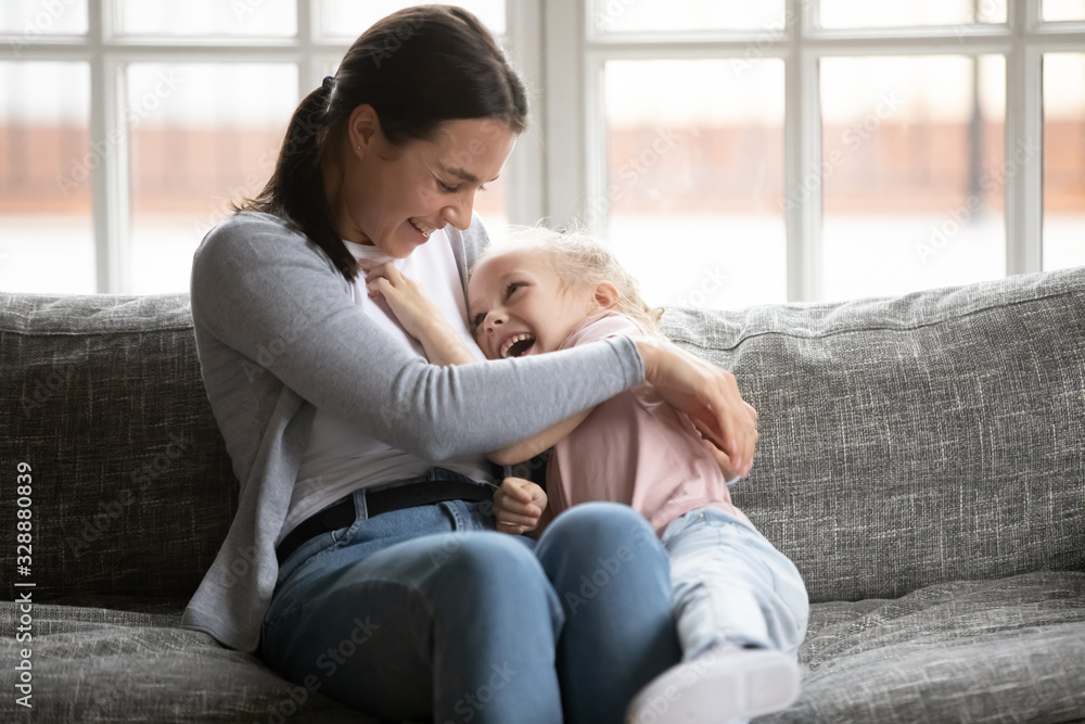 Happy young mother tickling little blonde daughter, having fun on comfortable sofa at home. Overjoyed small child girl playing with laughing nanny babysitter elder sister, enjoyin weekend time. Happy young mother tickling little blonde daughter, having fun on comfortable sofa at home. Overjoyed small child girl playing with laughing nanny babysitter elder sister, enjoyin weekend time.