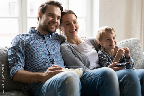 Smiling young man cuddling attractive wife and happy little preschool kid boy, relaxing on comfy sofa, watching show on tv. Joyful family couple enjoying leisure weekend time with small child at home.