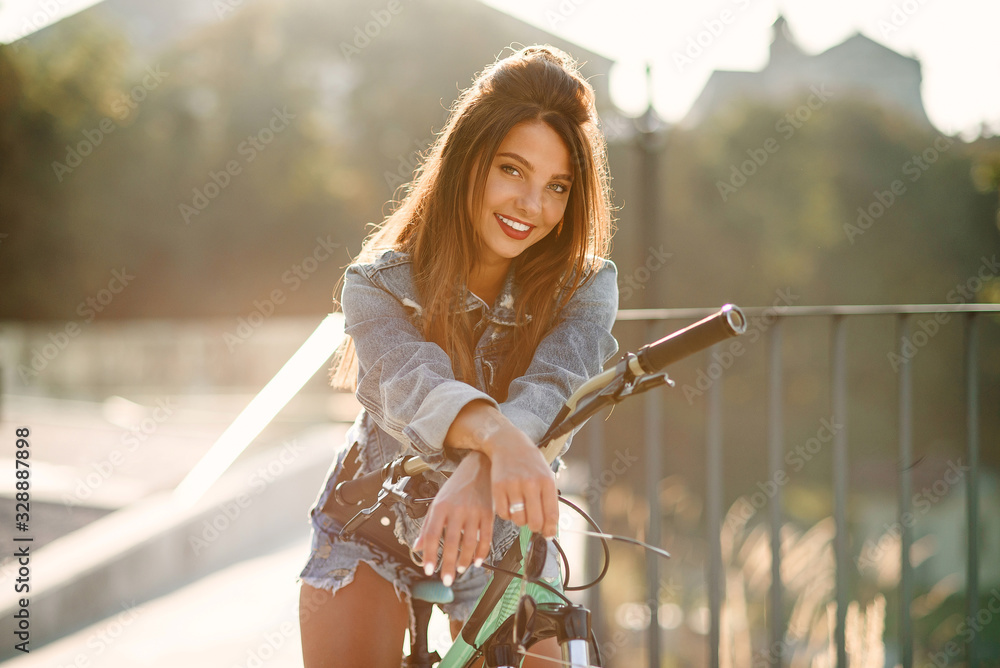 Attractive tempting young girl in fashion jeans clothes wears stylish glasses ,sitting on the bicycle and posing on camera at summer evening. Slow motion - obrazy, fototapety, plakaty 