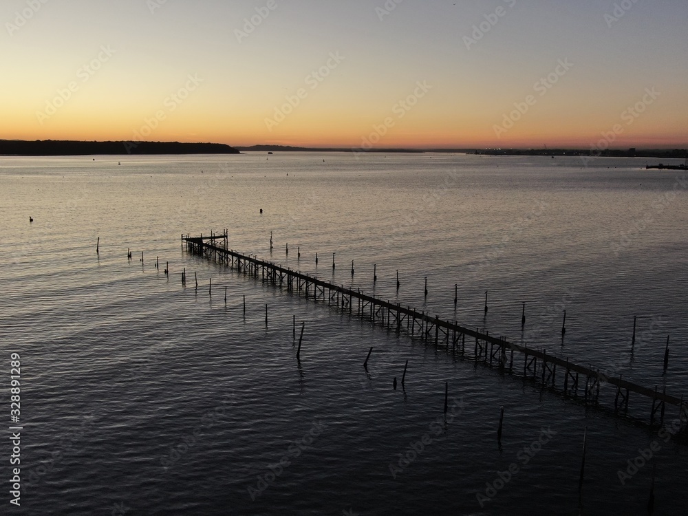 a wooden walkway or pier stretching diagonally into the sea at sunset with silhouetted islands in the background