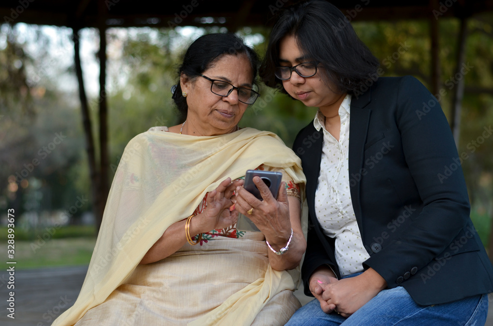 Smart young Indian girl with an old Indian woman looking at the mobile phone and busy talking sitting on a bench in a park in Delhi, India Smart young Indian girl with an old Indian woman looking at the mobile phone and busy talking sitting on a bench in a park in Delhi, India