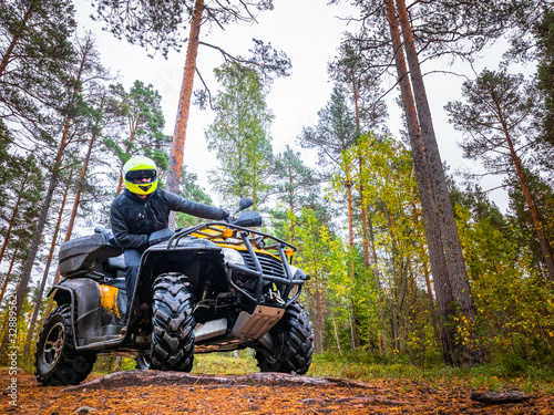 The driver of the ATV is looking at the camera. Man on a ATV in a yellow helmet. A trip through the forest on a ATV. Man is standing next to his qodracycle. Biker pans on the background of the forest