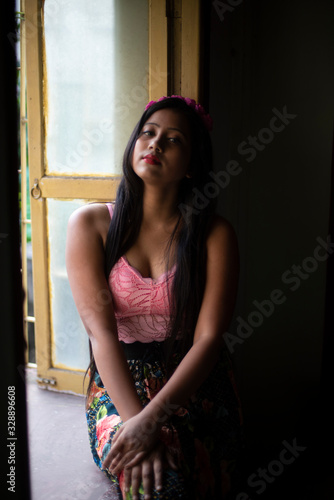 Portrait of an Asian/Japanese/Korean brunette young girl in pink top and skirt and floral headband in front of a window inside of a room. Fashion and boudoir photography. 