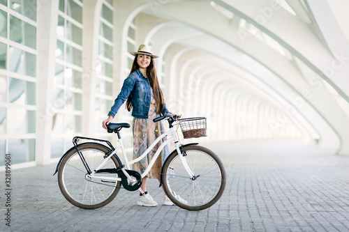 Fototapeta Naklejka Na Ścianę i Meble -  Teen girl with patterned dress and hat waits for a friend to go for a ride with her urban bicycle. Outdoor and fun concept in spring and summer