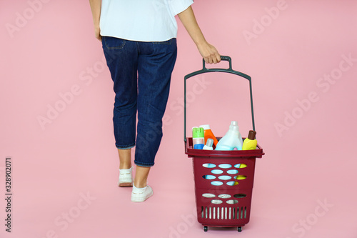 Woman with shopping basket full of cleaning supplies on pink background, closeup