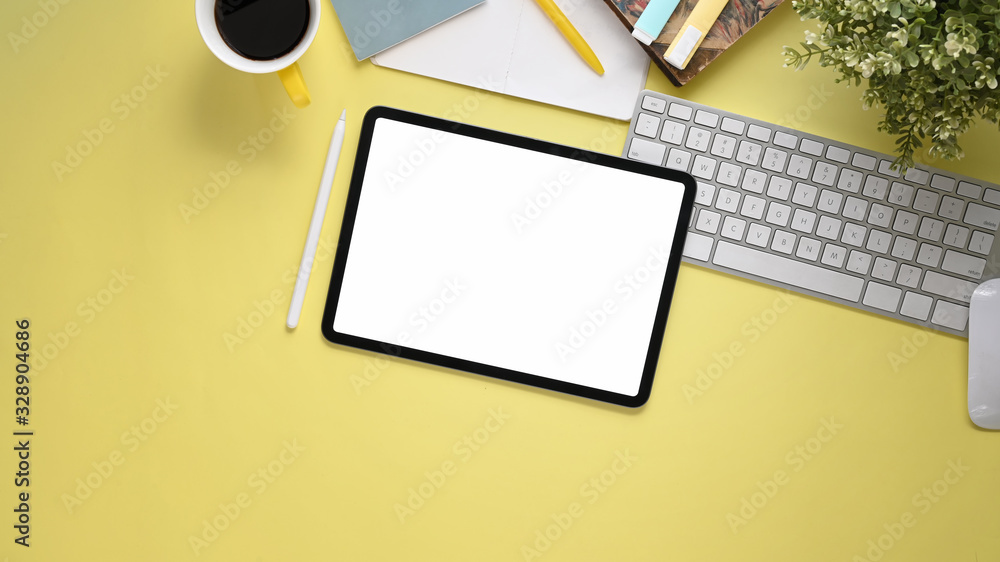 Top view image of workspace with flat lay white blank screen computer tablet, coffee cup, stylus pen, marker pens, stack of notebook, keyboard, mouse and potted plant on colorful working table. Top view image of workspace with flat lay white blank screen computer tablet, coffee cup, stylus pen, marker pens, stack of notebook, keyboard, mouse and potted plant on colorful working table.