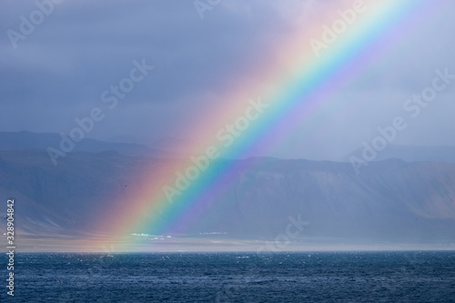 Beautiful summer landscape with sea  small village on the hill  bright colorful rainbow on blue sky  clouds and mountains on the horizon. Iceland  Peninsula